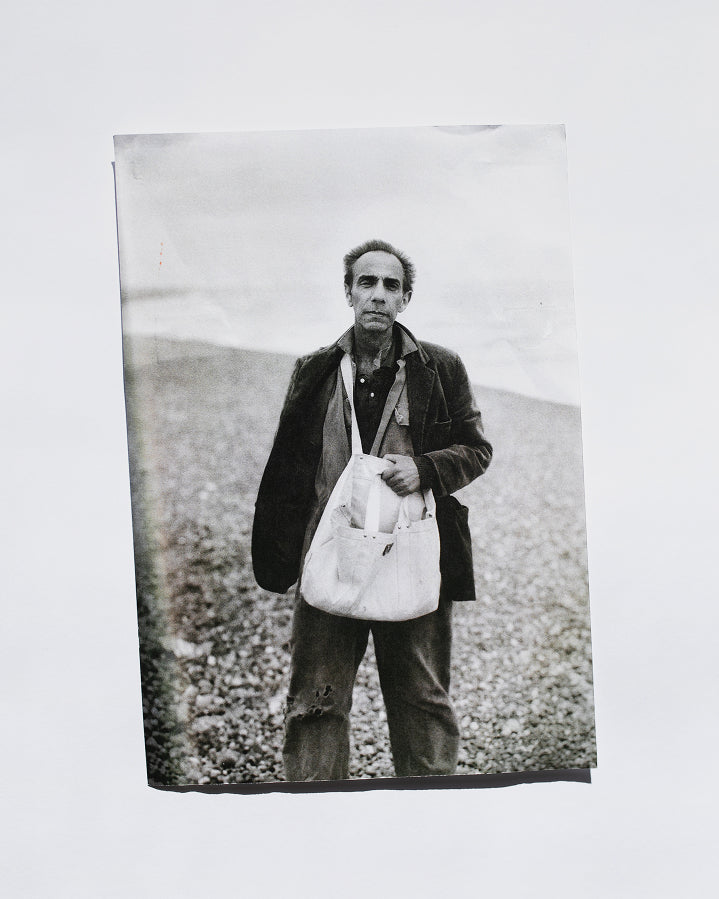 A black and white photographic print depicting a man standing on a pebble beach, holding a large white tote bag, shown against a plain white background.