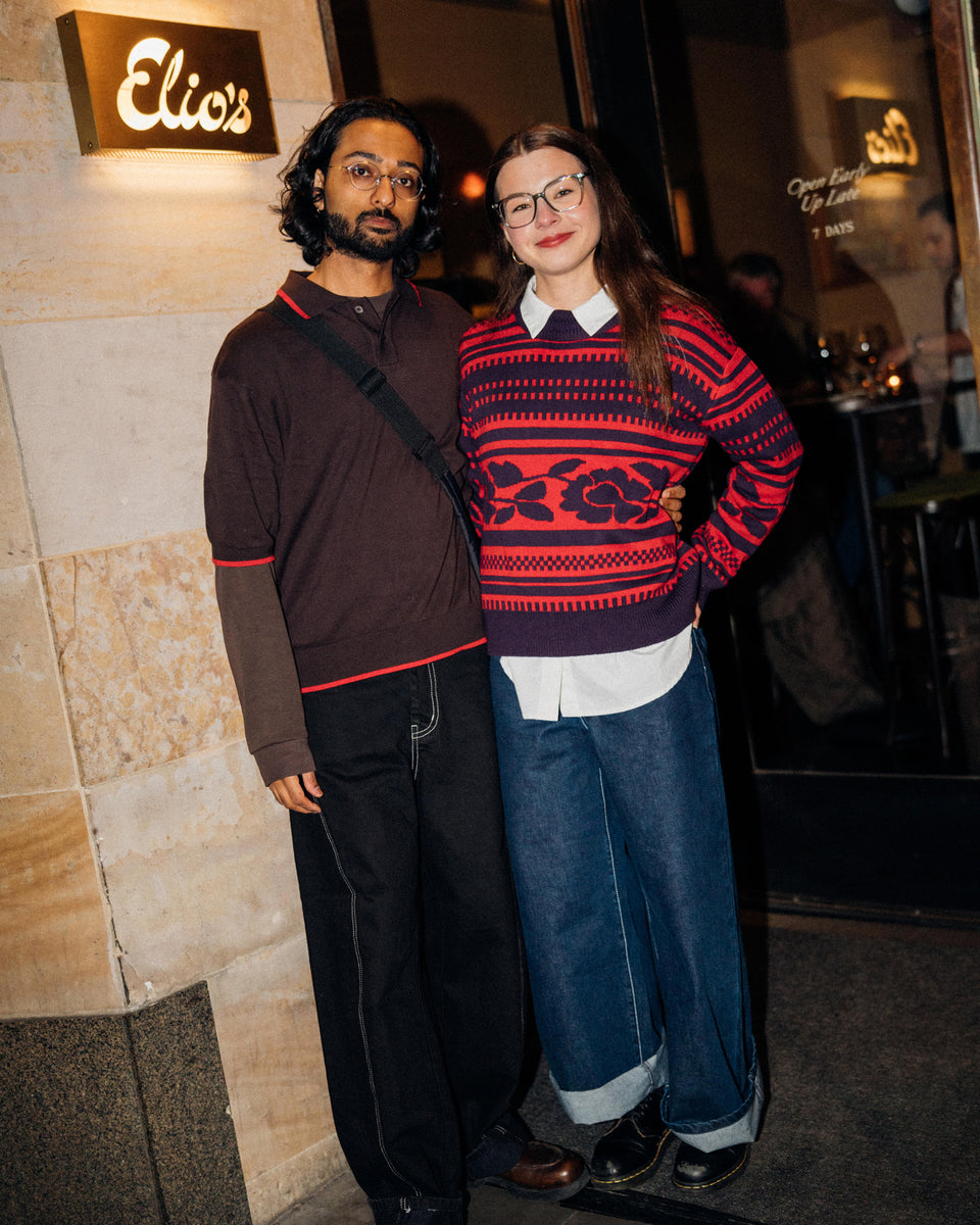 Two people posing together in front of a restaurant entrance. The woman wears a red and navy patterned knit sweater over a white collared shirt, blue jeans, and black shoes.