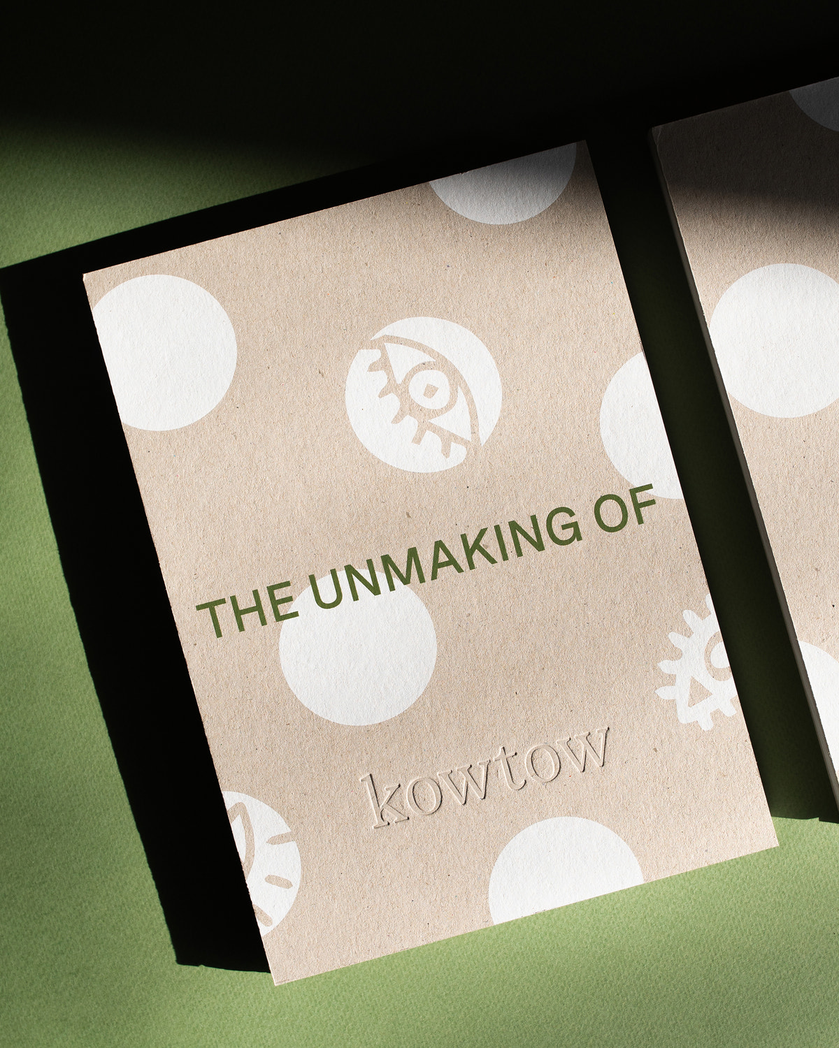 A close-up of a printed card or packaging piece with the text 'THE UNMAKING OF kowtow' on a kraft-paper style background with white circular shapes and embossed lettering, photographed on a green surface.