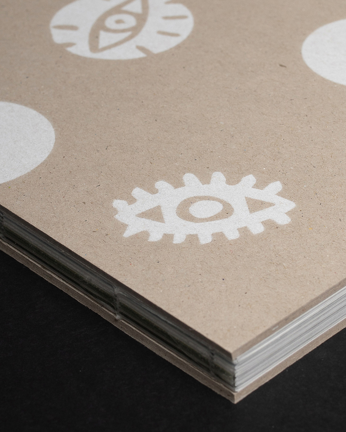A close-up of a kraft paper notebook or journal corner, showing white eye-and-gear style printed motifs on the cover, with multiple stacked pages visible along the edge, set against a dark background.