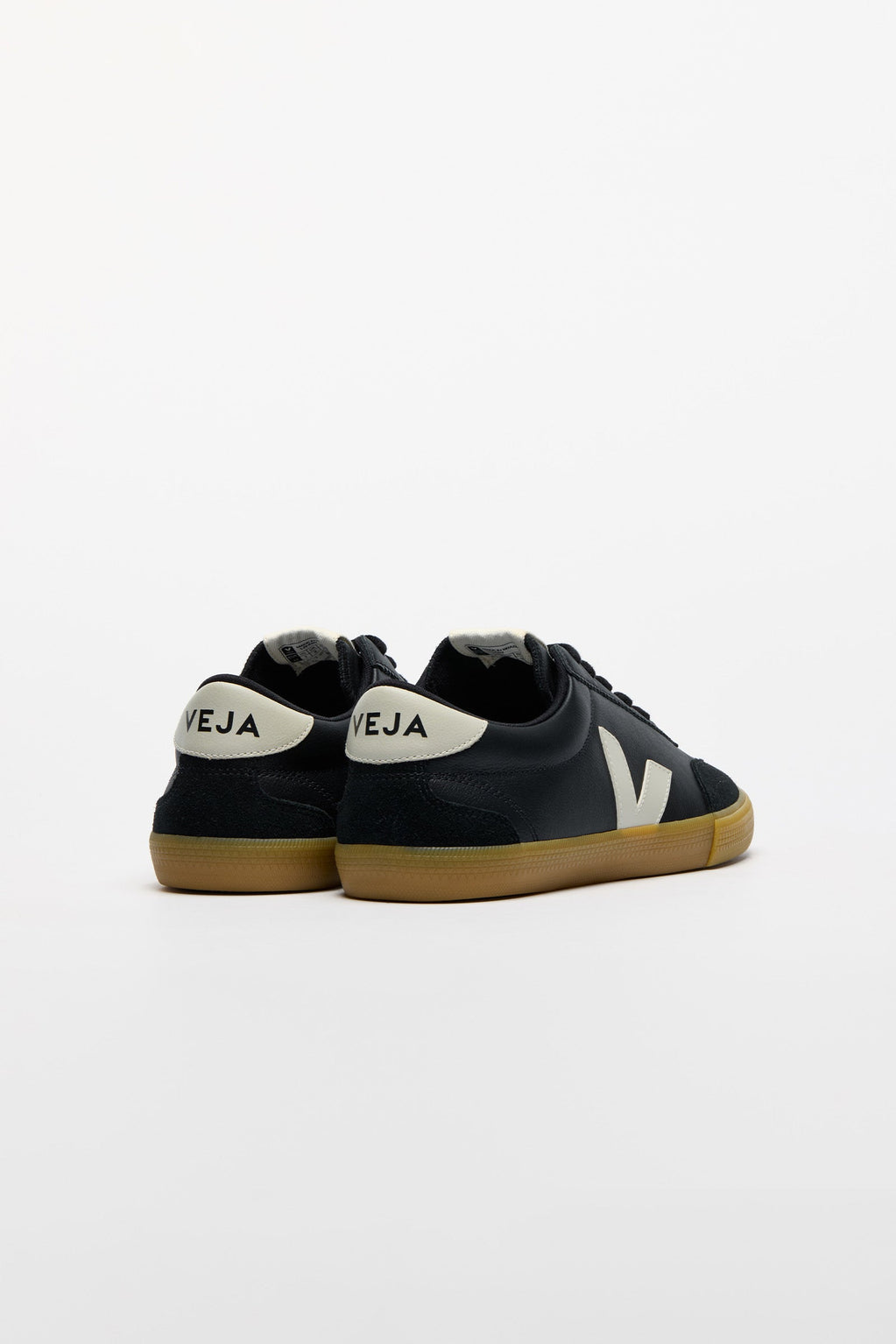 A pair of low-top sneakers shown from the back, featuring black suede uppers, a white heel tab with 'VEJA' printed in black, a white side logo, and a brown gum sole.