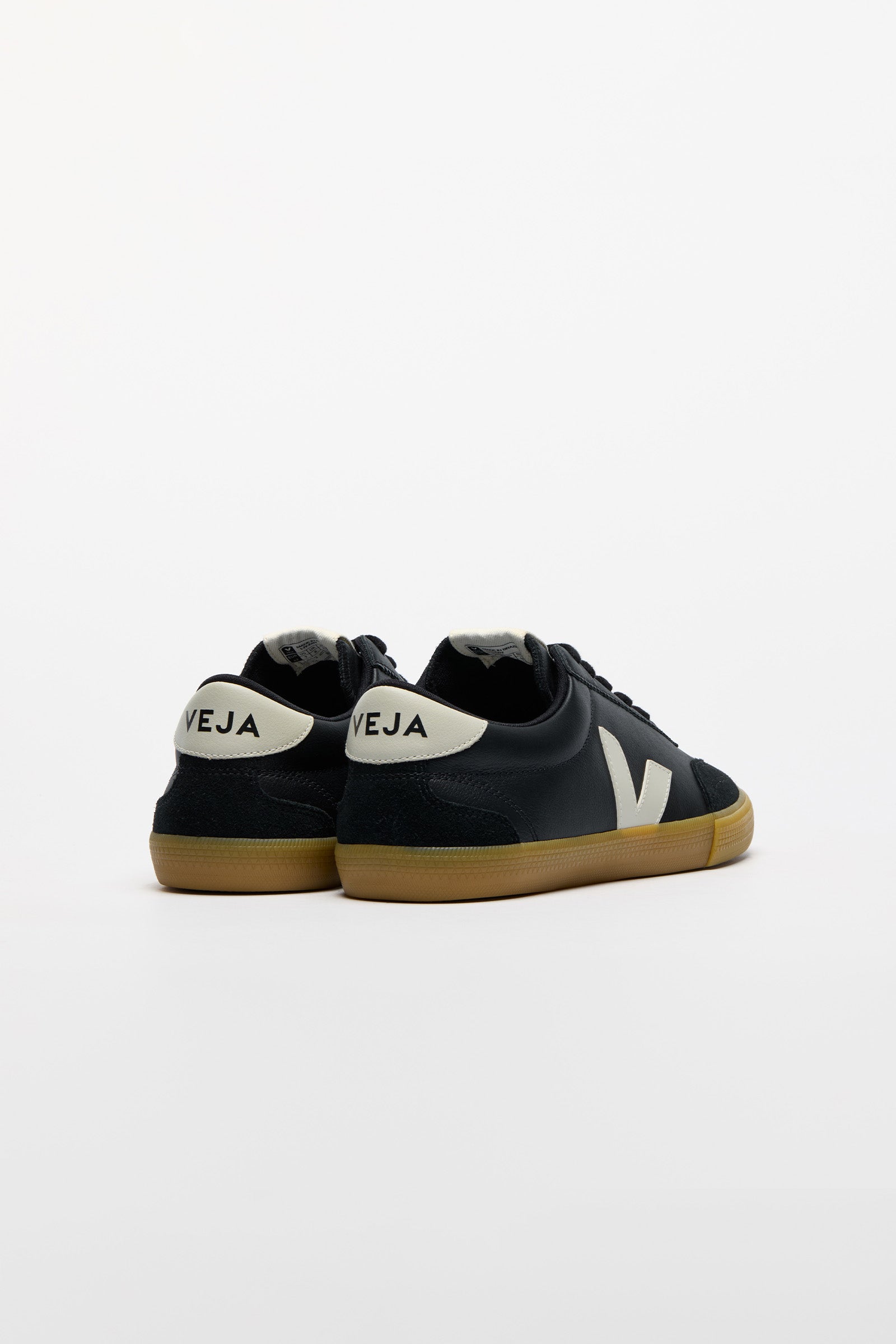 A pair of low-top sneakers shown from the back, featuring black suede uppers, a white heel tab with 'VEJA' printed in black, a white side logo, and a brown gum sole.