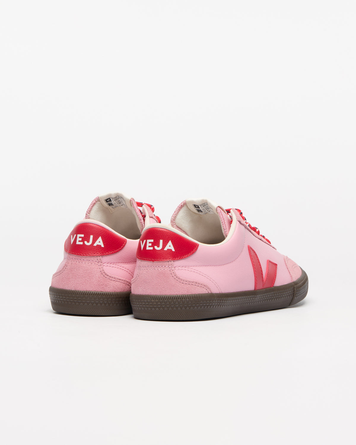 A pair of pink suede and leather sneakers with red accents, including a red heel tab with 'VEJA' in white text and a red side stripe.