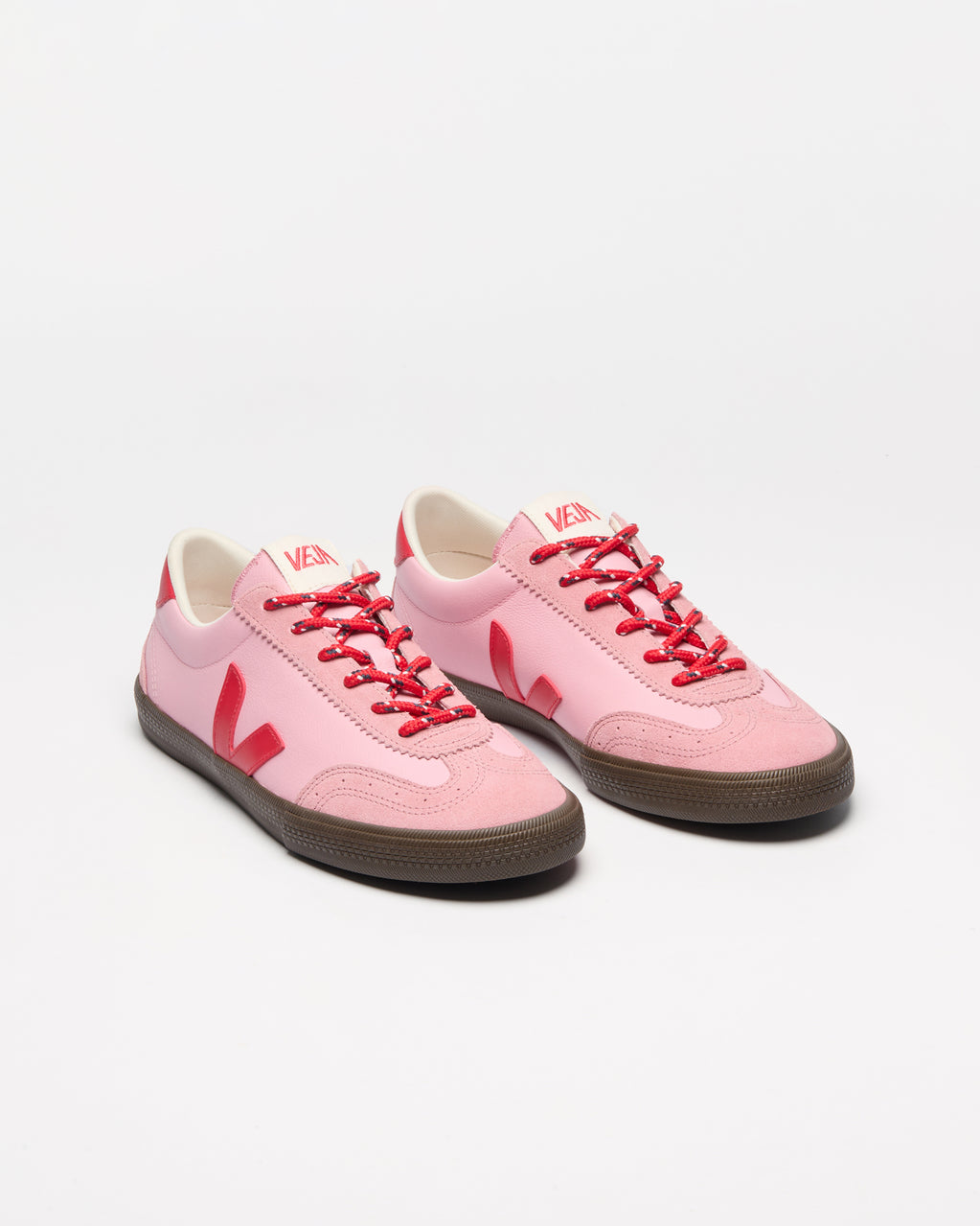 A pair of low-top sneakers in light pink with bright red laces, red logo accents, and dark brown rubber soles.