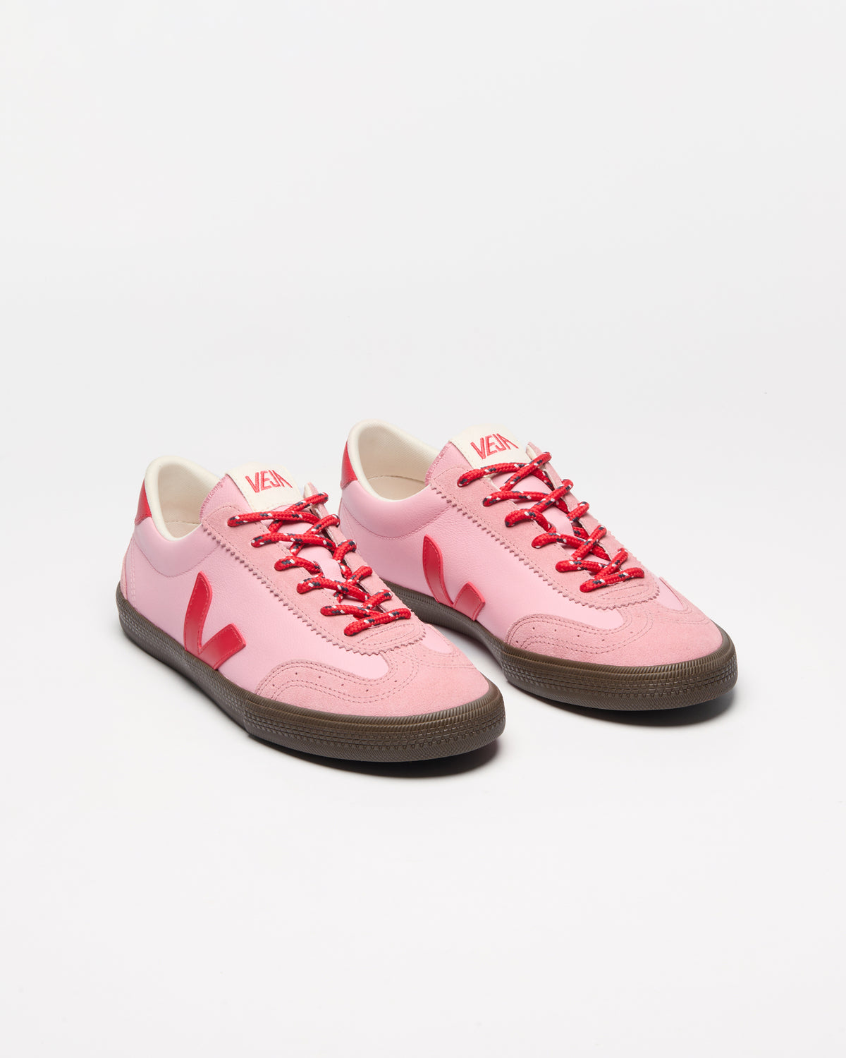 A pair of low-top sneakers in light pink with bright red laces, red logo accents, and dark brown rubber soles.