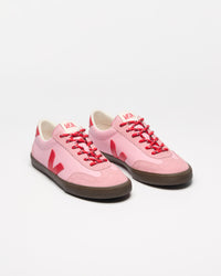A pair of low-top sneakers in light pink with bright red laces, red logo accents, and dark brown rubber soles.