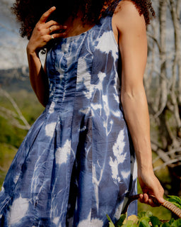 A woman outdoors wearing a sleeveless blue and white tie-dye dress, holding a basket of green leaves. The background shows blurred trees and outdoor scenery.
