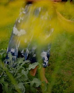 A person standing barefoot in a grassy outdoor garden, wearing a flowing blue and white tie-dye dress and holding a woven basket, with bright yellow flowers in the foreground.