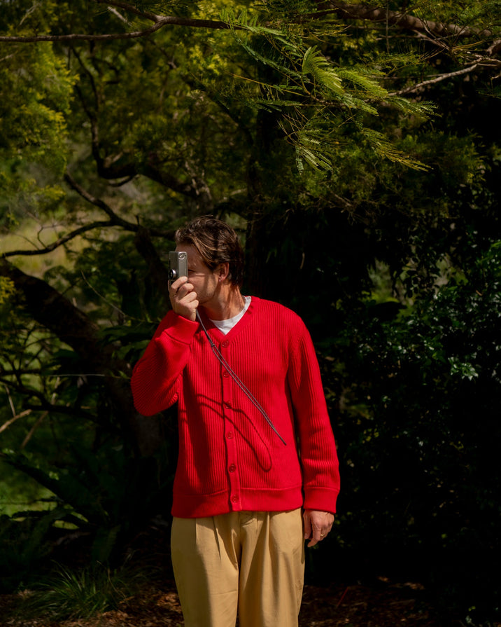 A person standing outdoors in a wooded area, wearing a bright red button up cardigan and beige pants, holding a clear camera to their face.