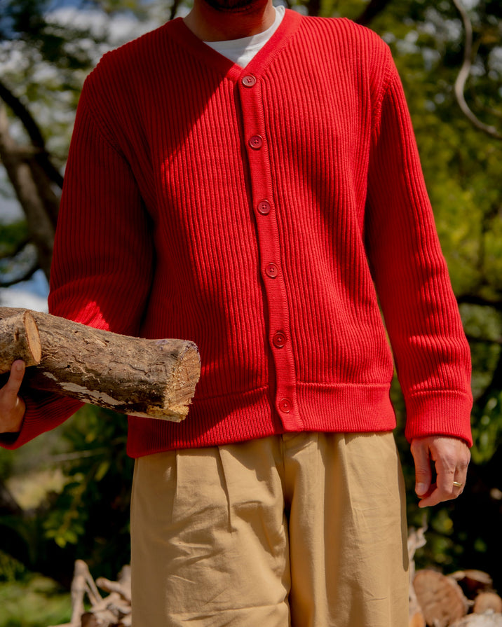 A person outdoors wearing a bright red ribbed button-front cardigan over a white undershirt and beige pants, holding a log. The background shows trees and stacked firewood in natural daylight.