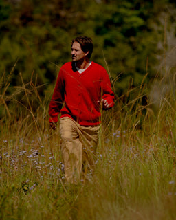 A man wearing a red knit cardigan and tan pants walking through a grassy outdoor field with trees in the background.