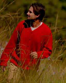 A man wearing a bright red knit cardigan over a white undershirt and tan pants, standing in tall sunlit grass outdoors.