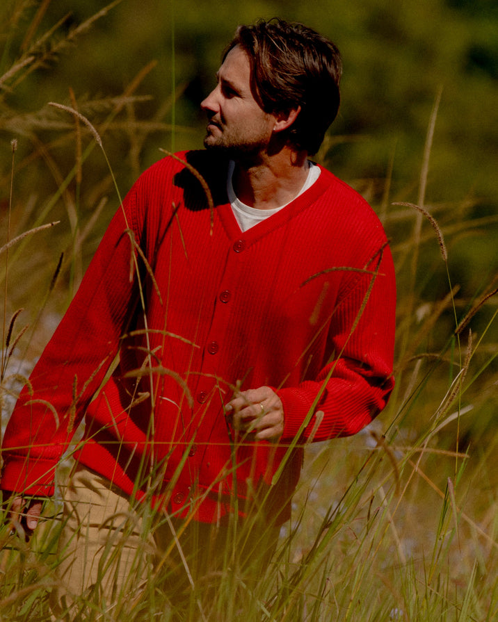 A man wearing a bright red knit cardigan over a white undershirt and tan pants, standing in tall sunlit grass outdoors.