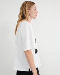 A woman shown in profile wearing an oversized white T-shirt with black graphic accents on the side, photographed against a plain light gray studio background.