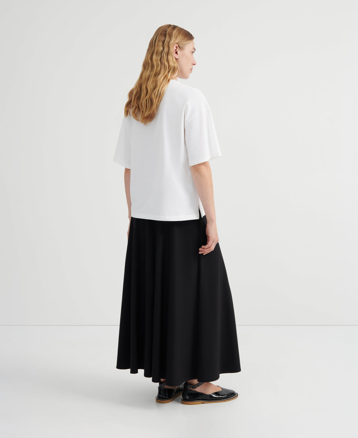 A woman shown from the back wearing a white short-sleeve top and a long black skirt, standing against a plain light gray studio background.
