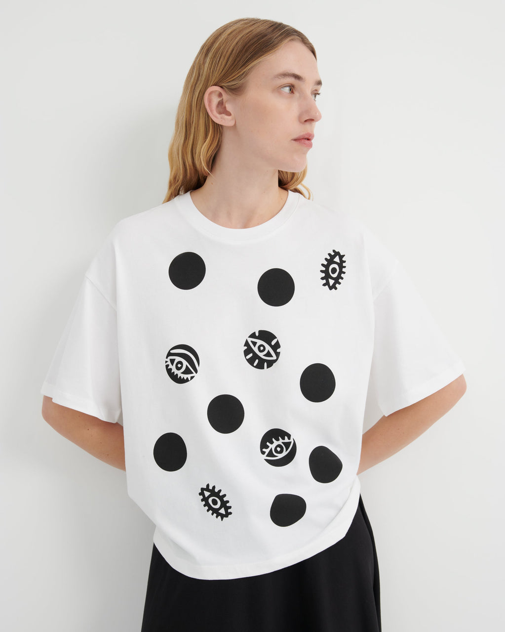 A woman wearing a white oversized T-shirt with black polka dots and eye-and-gear motifs, paired with black bottoms, photographed against a plain light gray studio background.