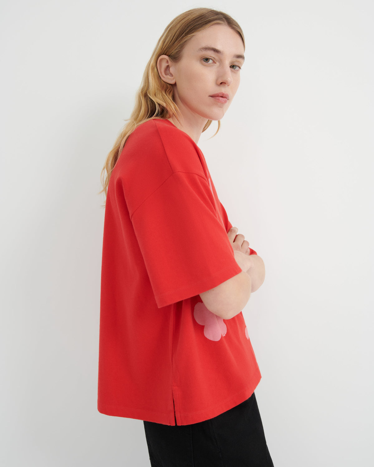 A woman wearing a bright red oversized short-sleeve top with a pink flower applique on the side, paired with black pants, photographed against a plain white studio background.