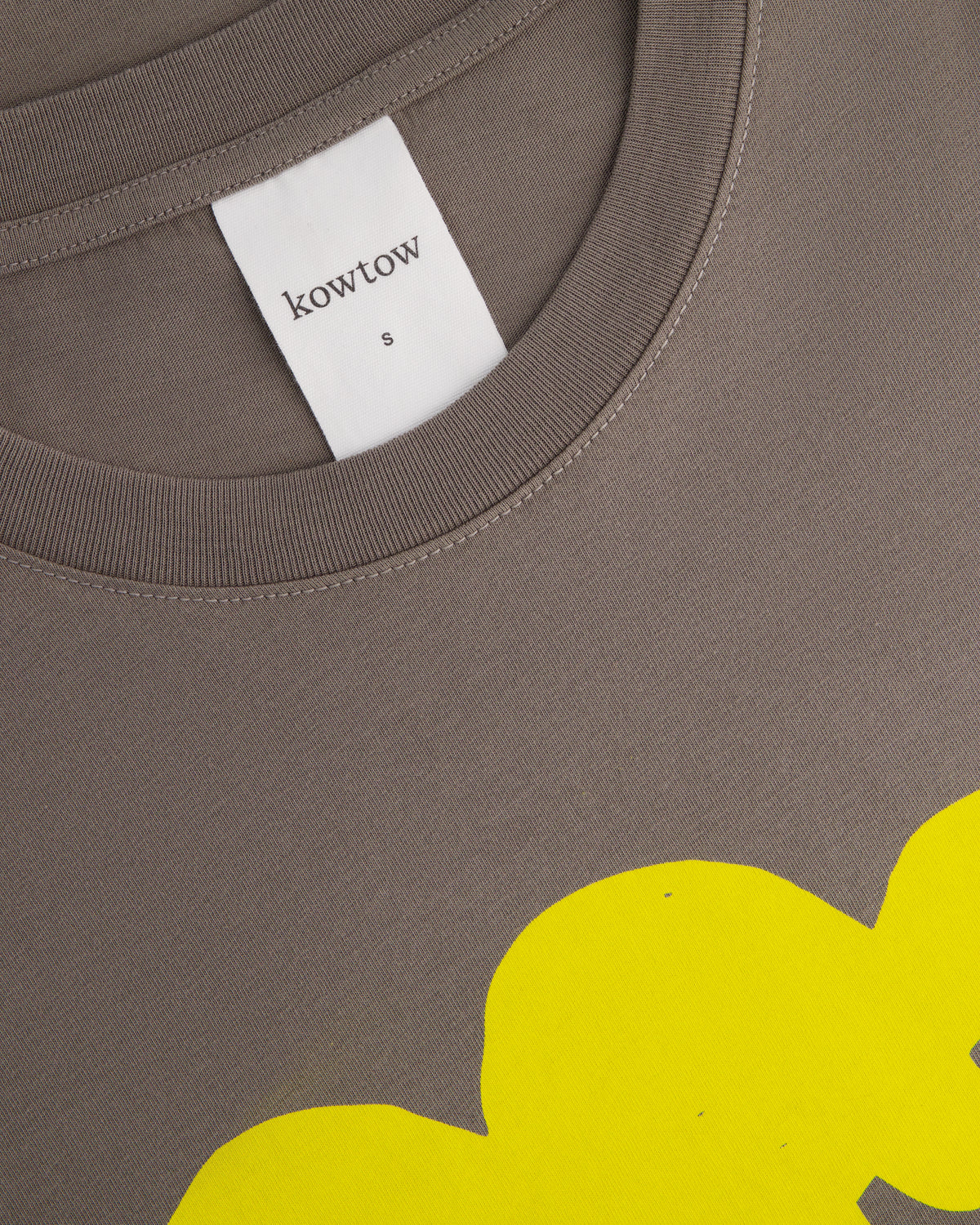 Close-up of a Kowtow brand crewneck sweatshirt in a muted taupe-brown color, featuring a white neck label and a bright yellow graphic print at the bottom.
