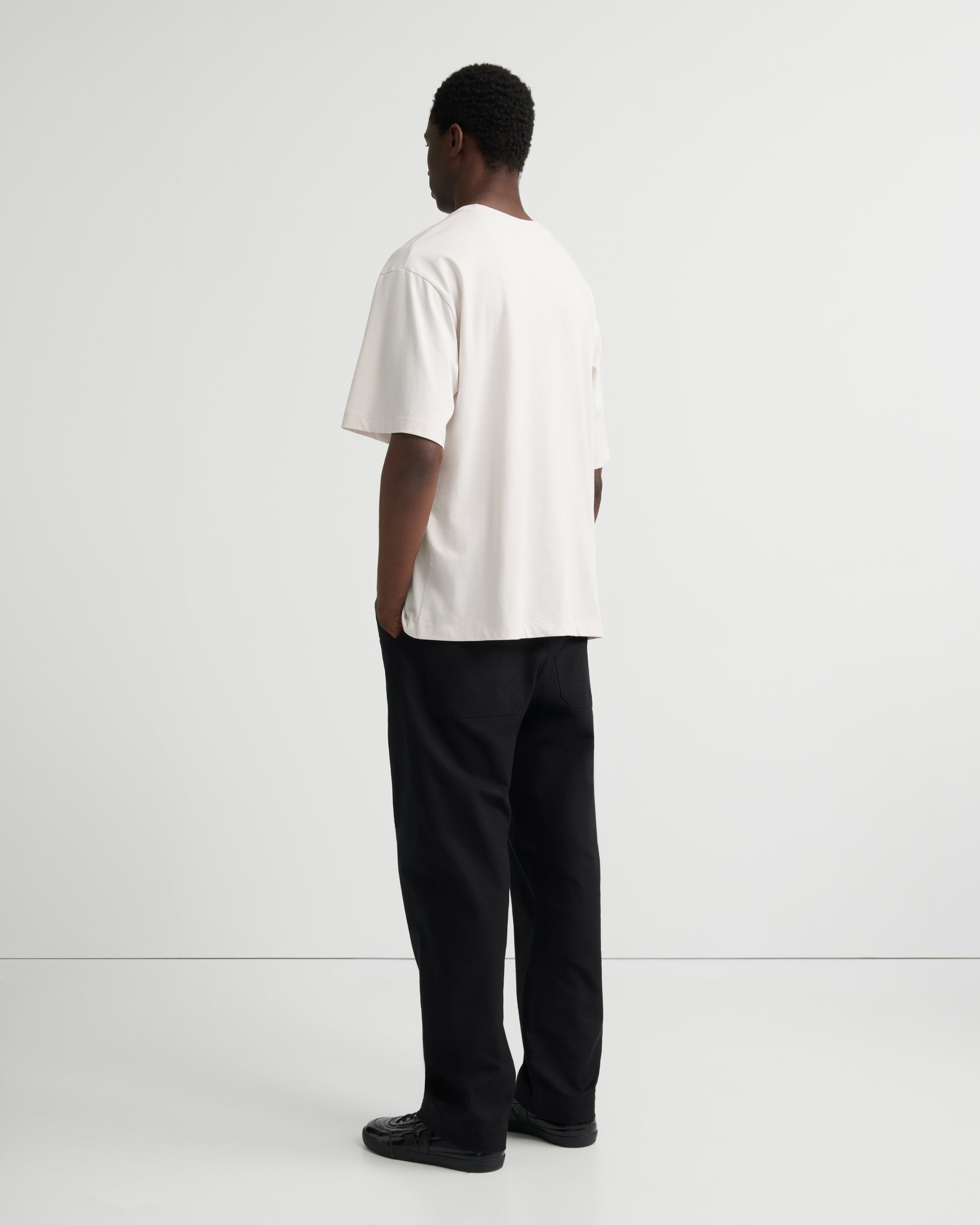 A man standing with his back to the camera, wearing a loose off-white short-sleeve t-shirt and black trousers.