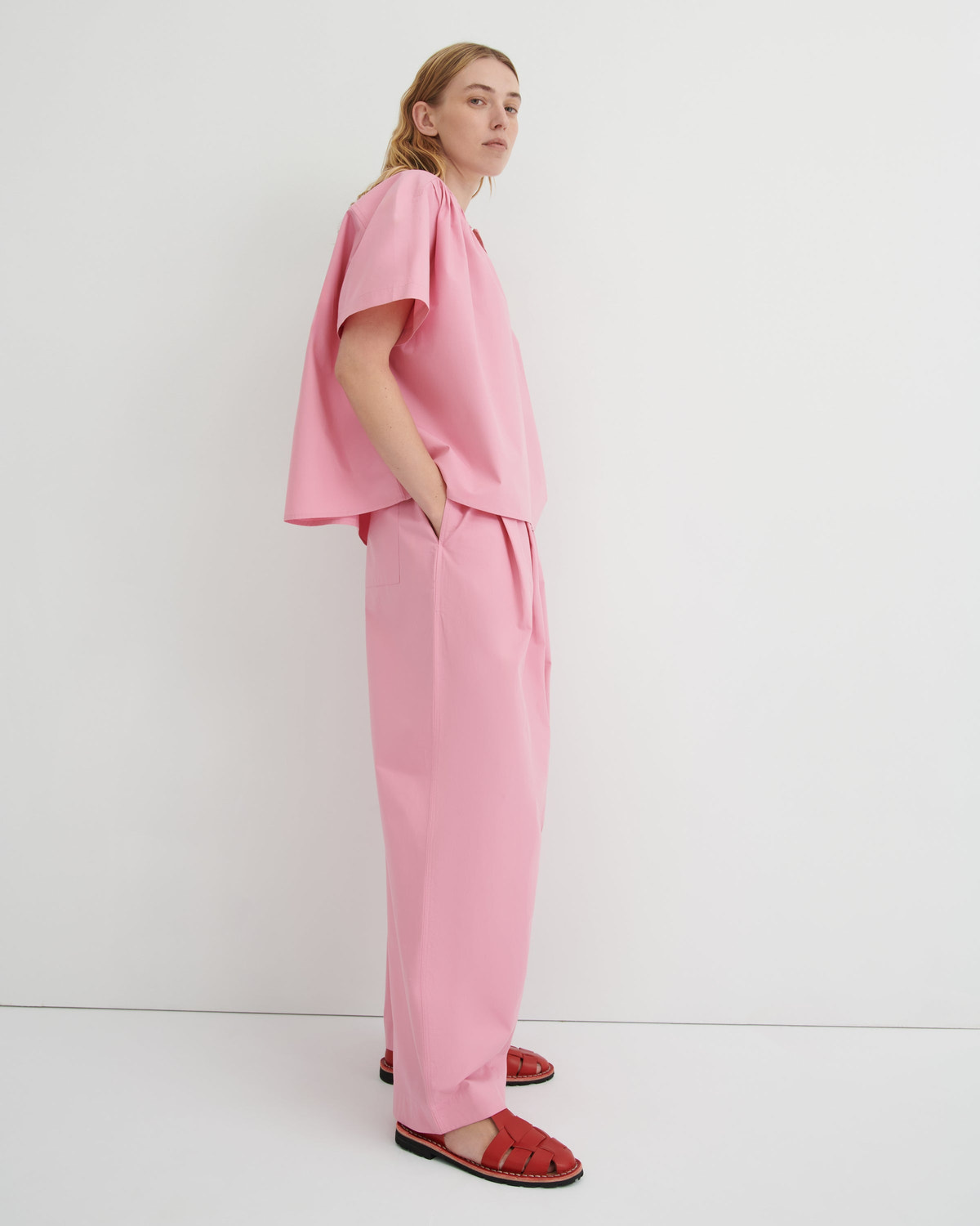 A woman stands in profile wearing a matching light pink two-piece outfit with a short flutter-sleeve top and wide-leg trousers, paired with red sandals, against a plain white studio background.