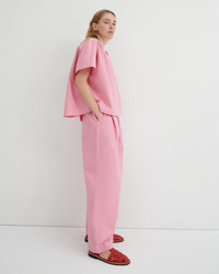 A woman stands in profile wearing a matching light pink two-piece outfit with a short flutter-sleeve top and wide-leg trousers, paired with red sandals, against a plain white studio background.