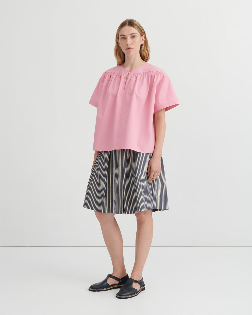 A woman modeling a light pink short-sleeve blouse with a gathered neckline and pleated skirt in a studio setting.