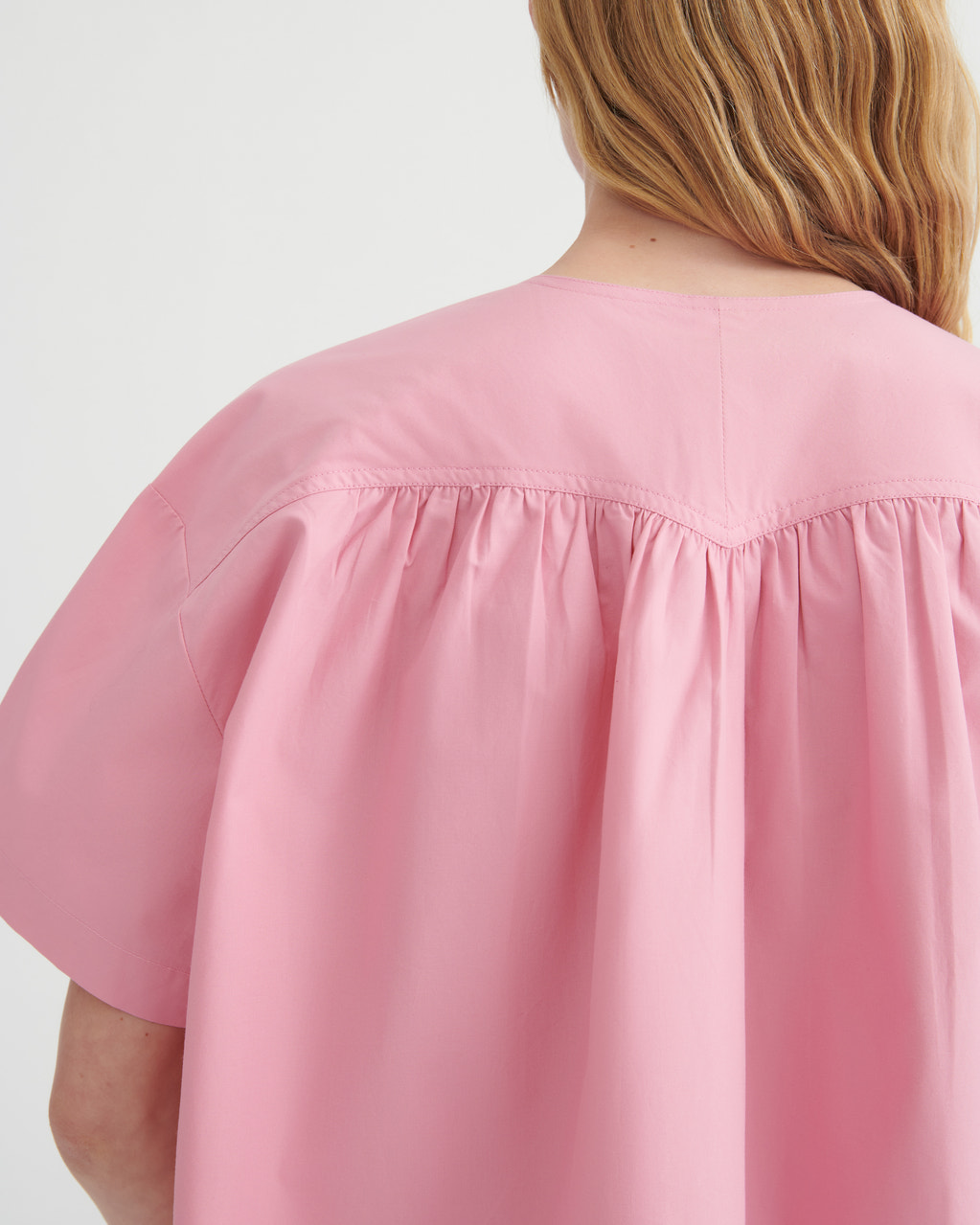 Close-up back view of a person wearing a loose pink short-sleeve top with gathered shoulder seams, against a plain light background.