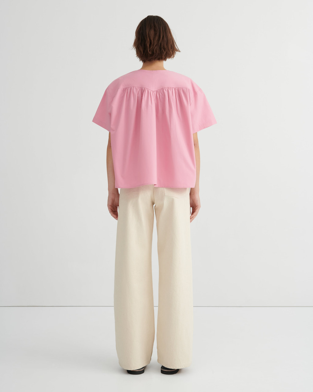 A woman stands backwards wearing a light pink short-sleeve blouse with a gathered neckline and wide-leg cream trousers, paired with red sandals, against a clean white studio background.