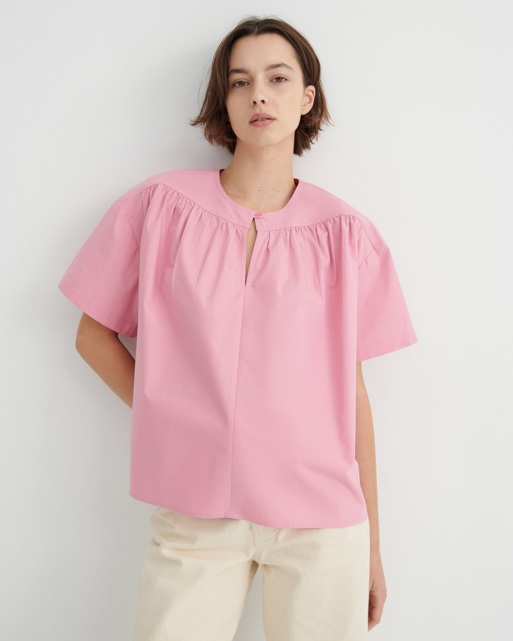 A woman stands in profile wearing a light pink short-sleeve blouse with a gathered neckline.