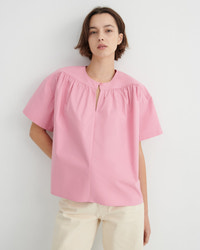 A woman stands in profile wearing a light pink short-sleeve blouse with a gathered neckline.