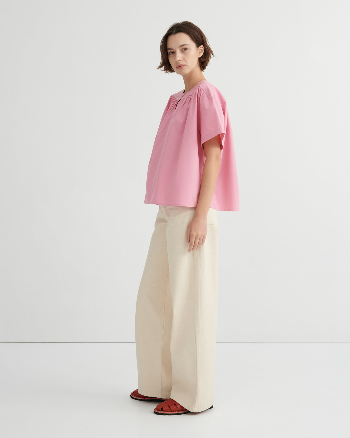 A woman stands in profile wearing a light pink short-sleeve blouse with a gathered neckline and wide-leg cream trousers, paired with red sandals, against a clean white studio background.