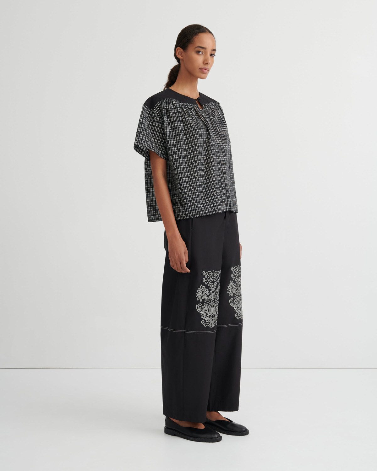 A model stands in a studio wearing a short-sleeve black and white checkered top paired with wide-leg black trousers featuring white floral embroidery near the lower leg, and black flat shoes.