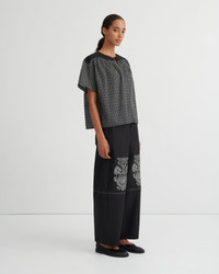 A model stands in a studio wearing a short-sleeve black and white checkered top paired with wide-leg black trousers featuring white floral embroidery near the lower leg, and black flat shoes.