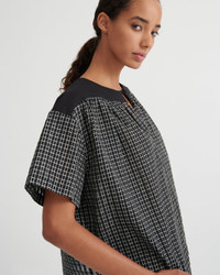A woman wearing a loose short-sleeve top with a black and white grid pattern and solid black shoulder yoke, photographed against a light gray studio background.