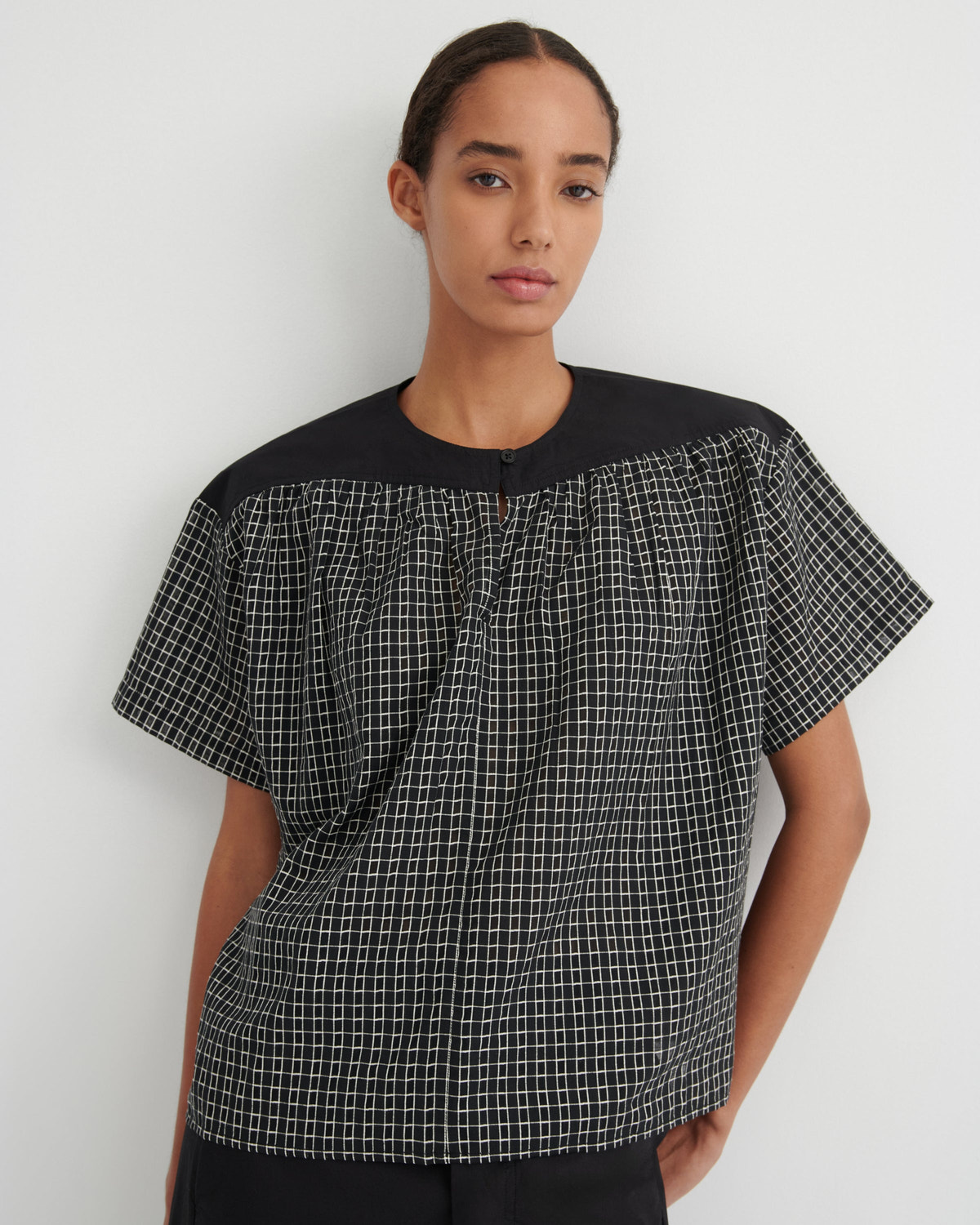A woman modeling a short-sleeve top with a black and white grid pattern on the body and solid black shoulders and neckline, set against a plain light gray background.