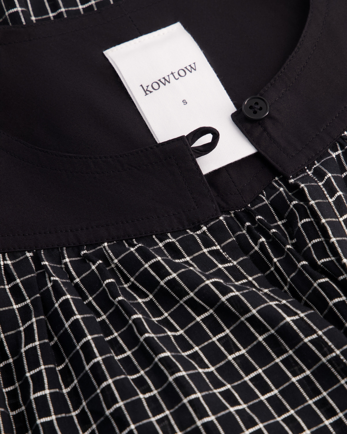 Close-up of a Kowtow garment showing a black collar with a white brand tag, a black button, and a black-and-white grid patterned fabric below.