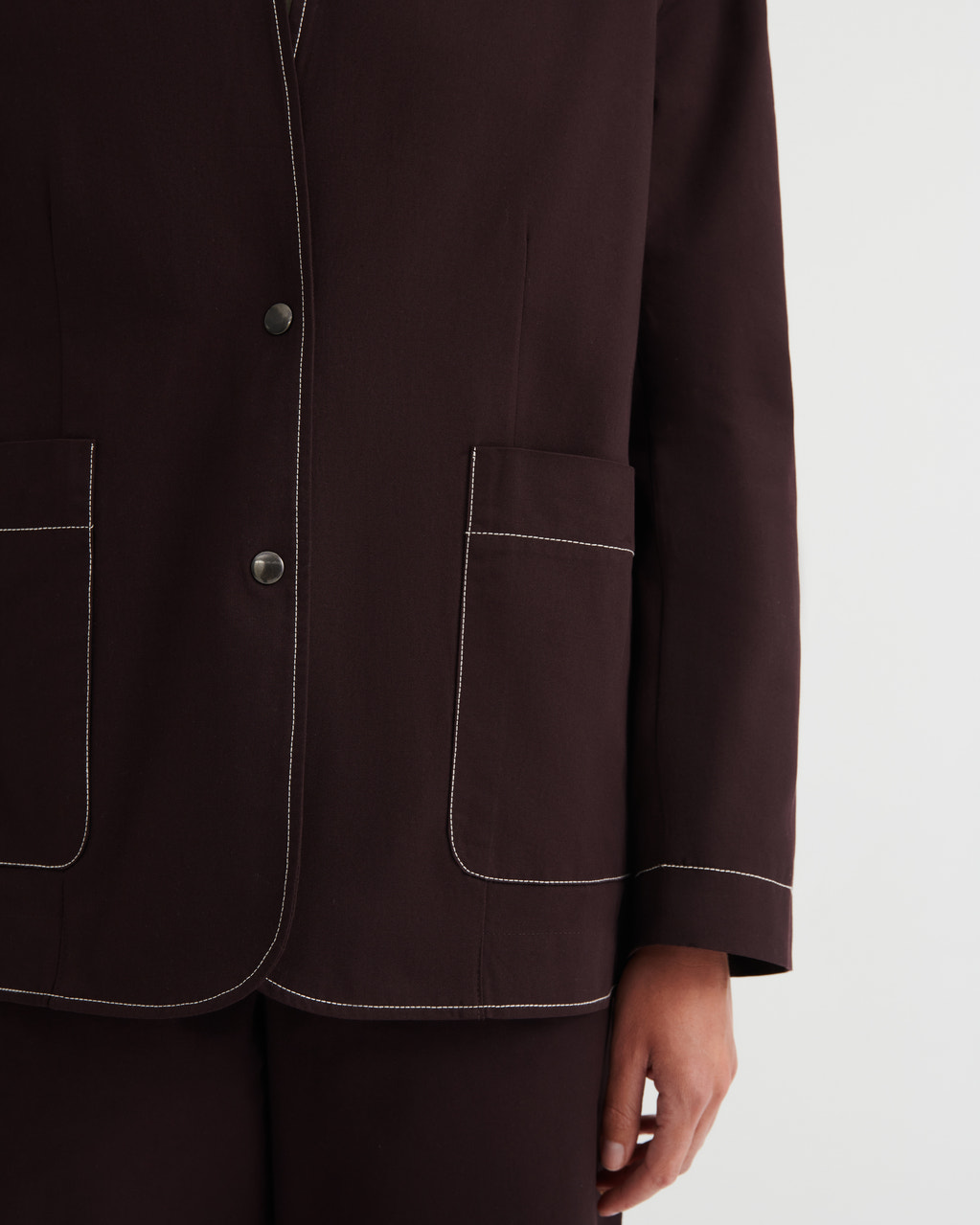 Close-up of a dark brown tailored jacket with white contrast stitching, featuring two large front patch pockets and a button closure, worn by a model against a light gray background.
