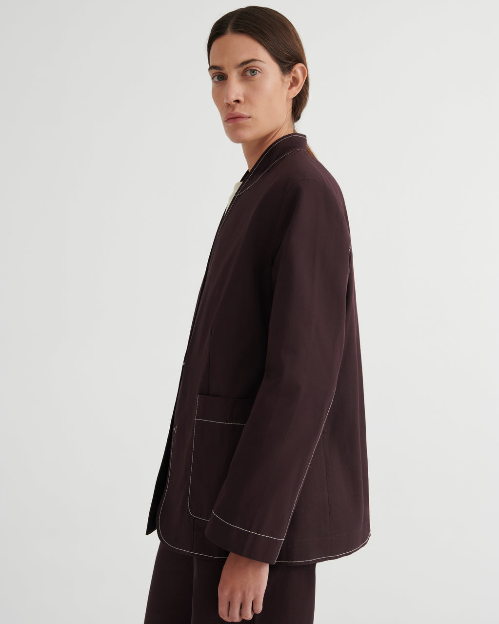 A model shown from the side in a dark brown blazer with contrasting light piping, paired with matching dark trousers, against a plain light gray studio background.