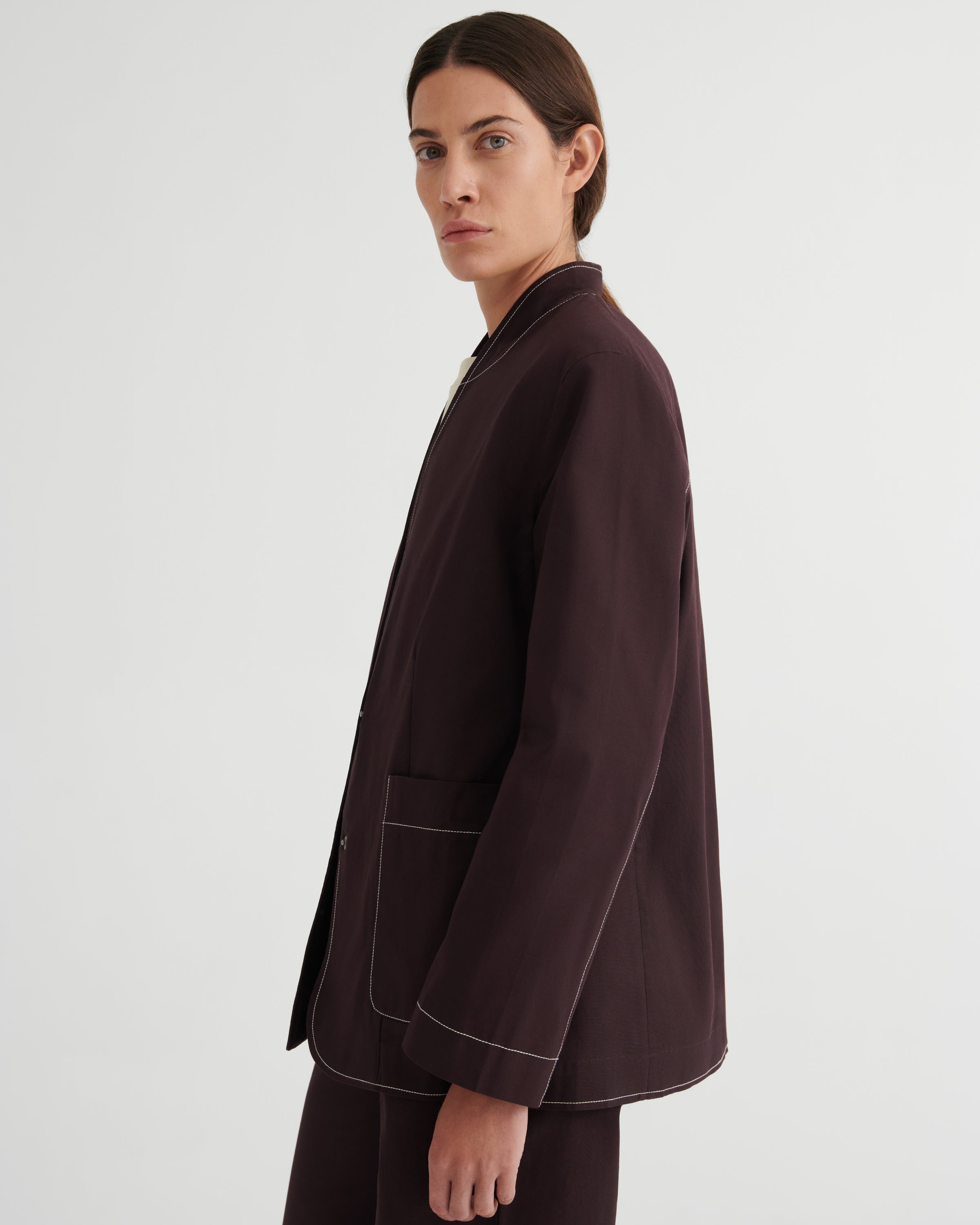 A model shown from the side in a dark brown blazer with contrasting light piping, paired with matching dark trousers, against a plain light gray studio background.