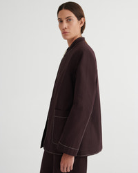 A model shown from the side in a dark brown blazer with contrasting light piping, paired with matching dark trousers, against a plain light gray studio background.