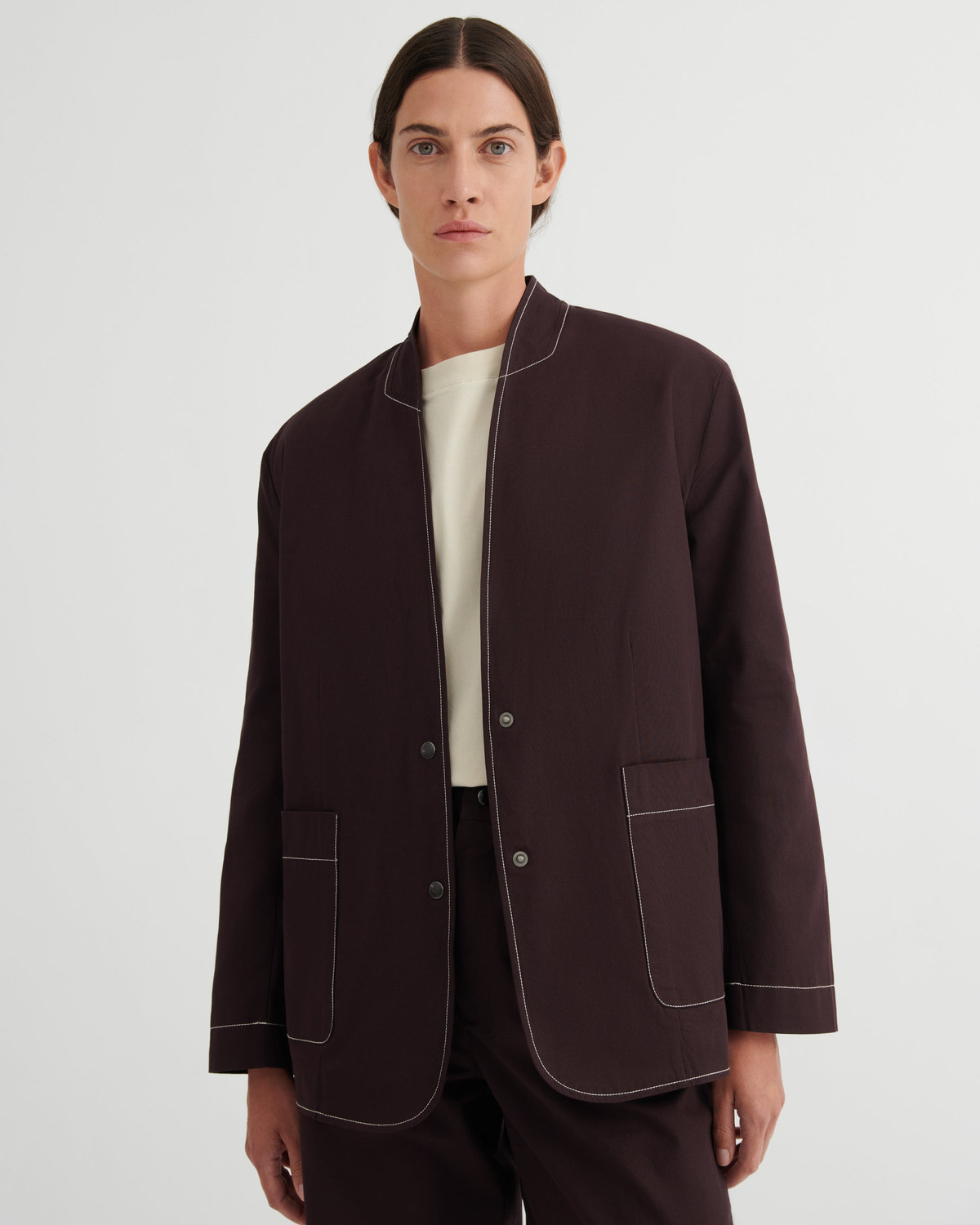A model wearing a dark brown jacket with contrasting light beige piping, styled over a cream shirt and dark trousers, shot against a light gray studio background.