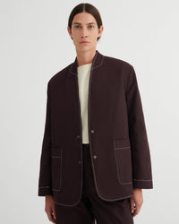 A model wearing a dark brown jacket with contrasting light beige piping, styled over a cream shirt and dark trousers, shot against a light gray studio background.