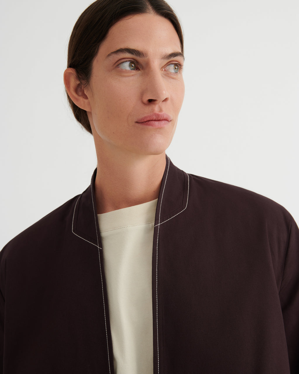 A close-up of a model wearing a dark brown bomber jacket with white contrast stitching over a cream crewneck shirt, set against a plain light gray background.