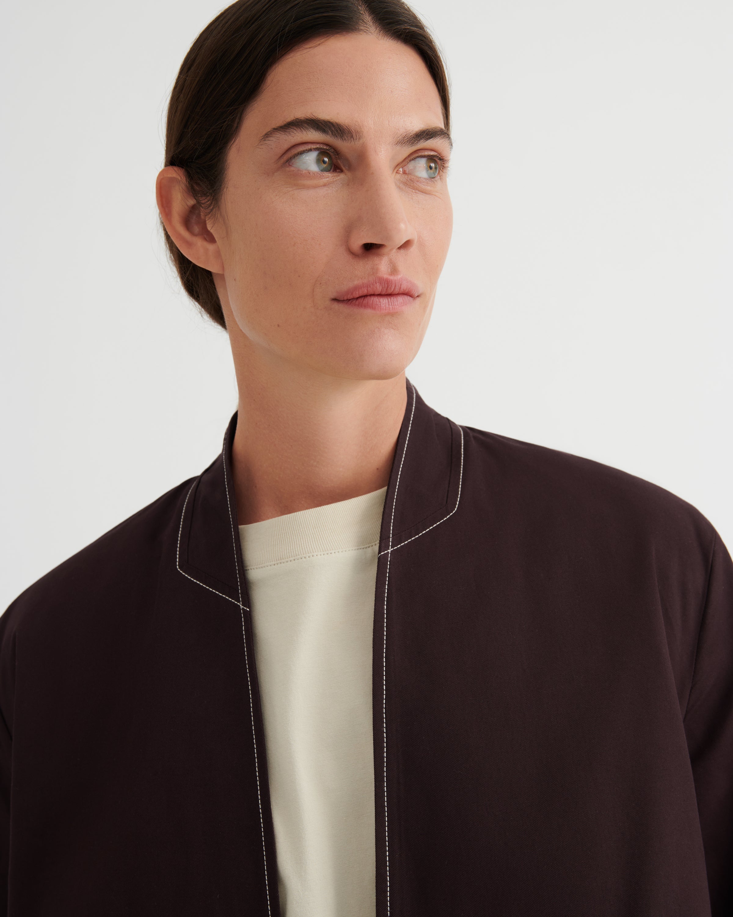 A close-up of a model wearing a dark brown bomber jacket with white contrast stitching over a cream crewneck shirt, set against a plain light gray background.