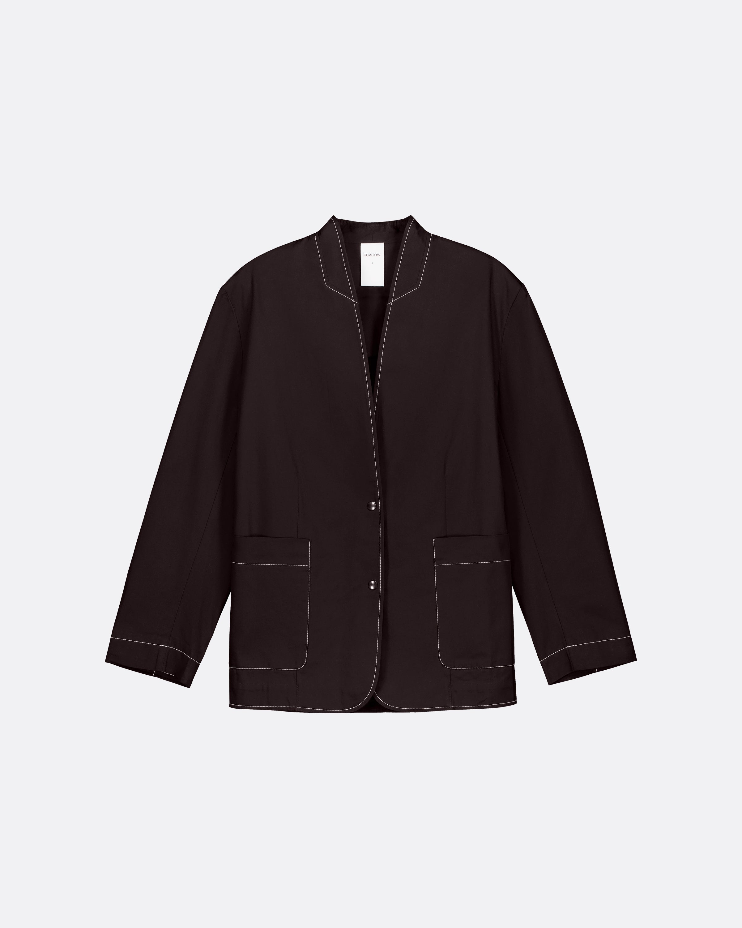 A dark brown cropped jacket with white contrast stitching, featuring a stand collar, button front, and two front patch pockets, displayed on a light gray background.