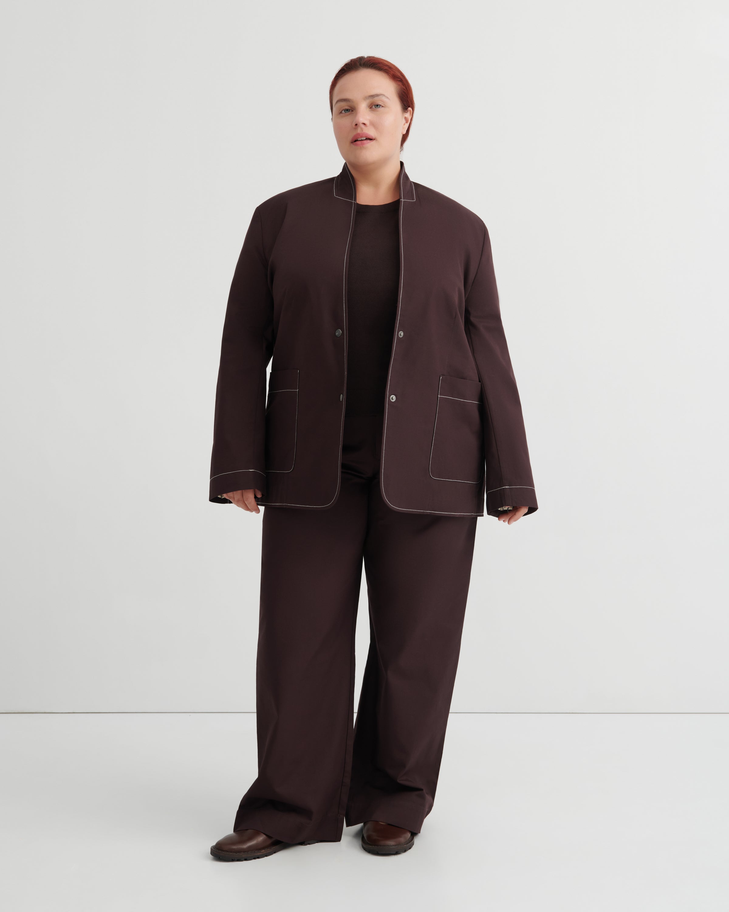 A model stands against a light gray studio background wearing a dark brown matching set consisting of a cropped jacket with contrast light piping, wide-leg trousers, and a black top.