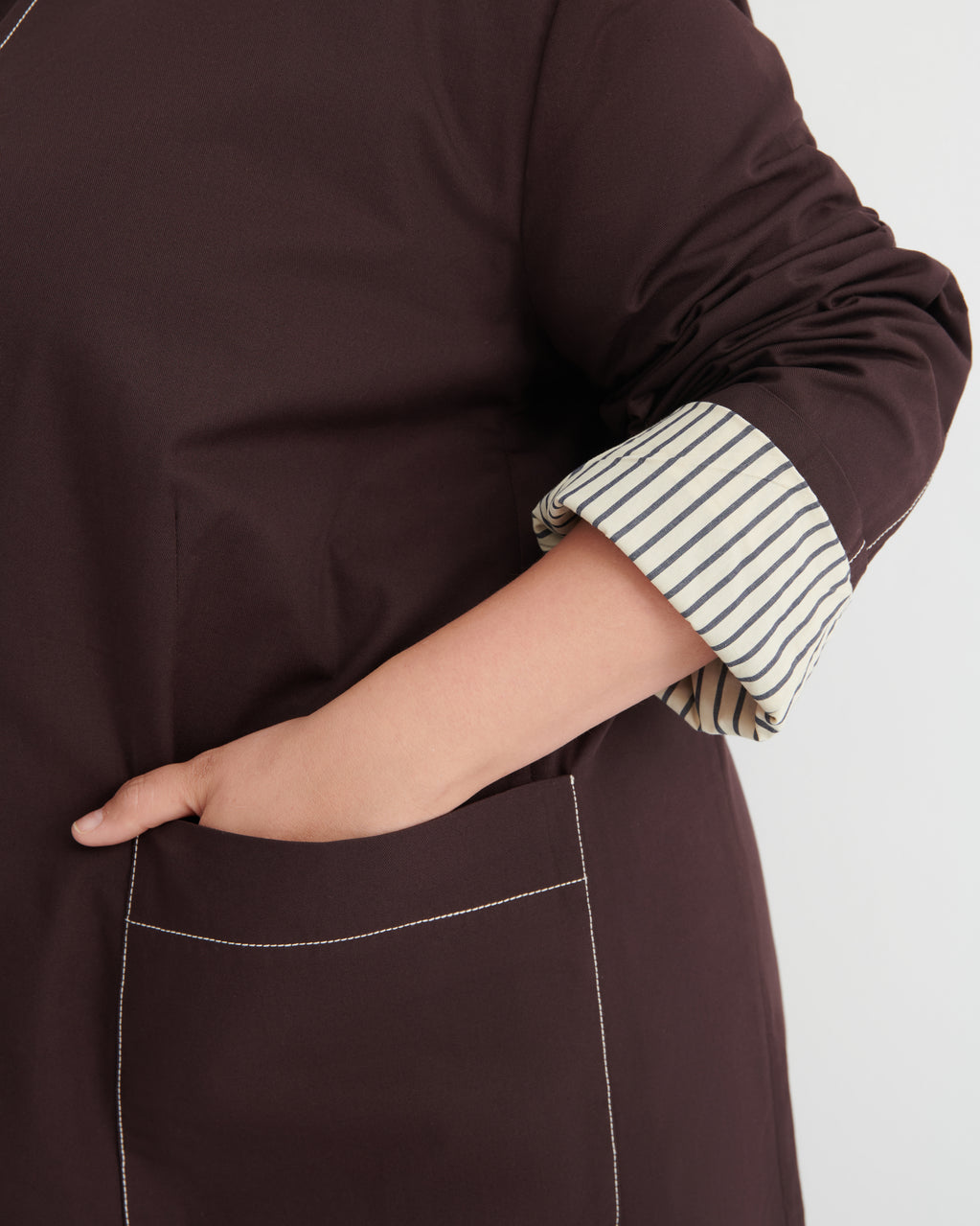 A close-up of a person wearing a dark brown long-sleeve top or dress with a striped button-up shirt rolled up at the sleeve, showing a hand in a pocket against a light gray background.