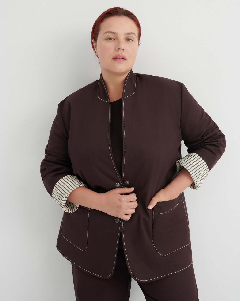 A woman modeling a dark brown jacket with contrasting light topstitching and striped button-up cuffs, paired with matching dark brown trousers against a light gray studio background.