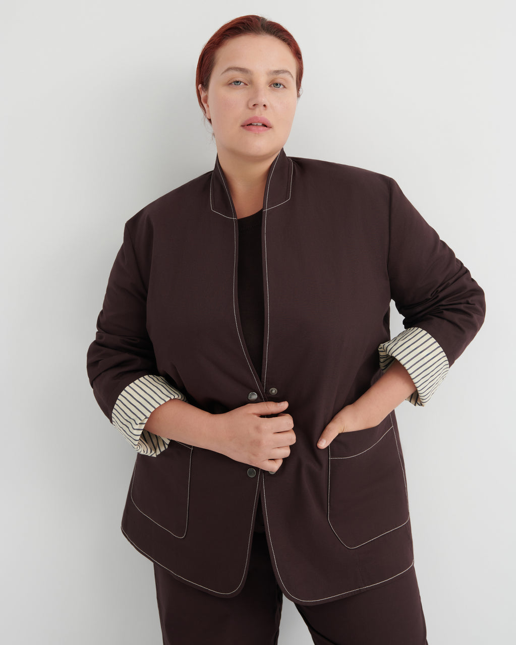 A woman modeling a dark brown jacket with contrasting light topstitching and striped button-up cuffs, paired with matching dark brown trousers against a light gray studio background.