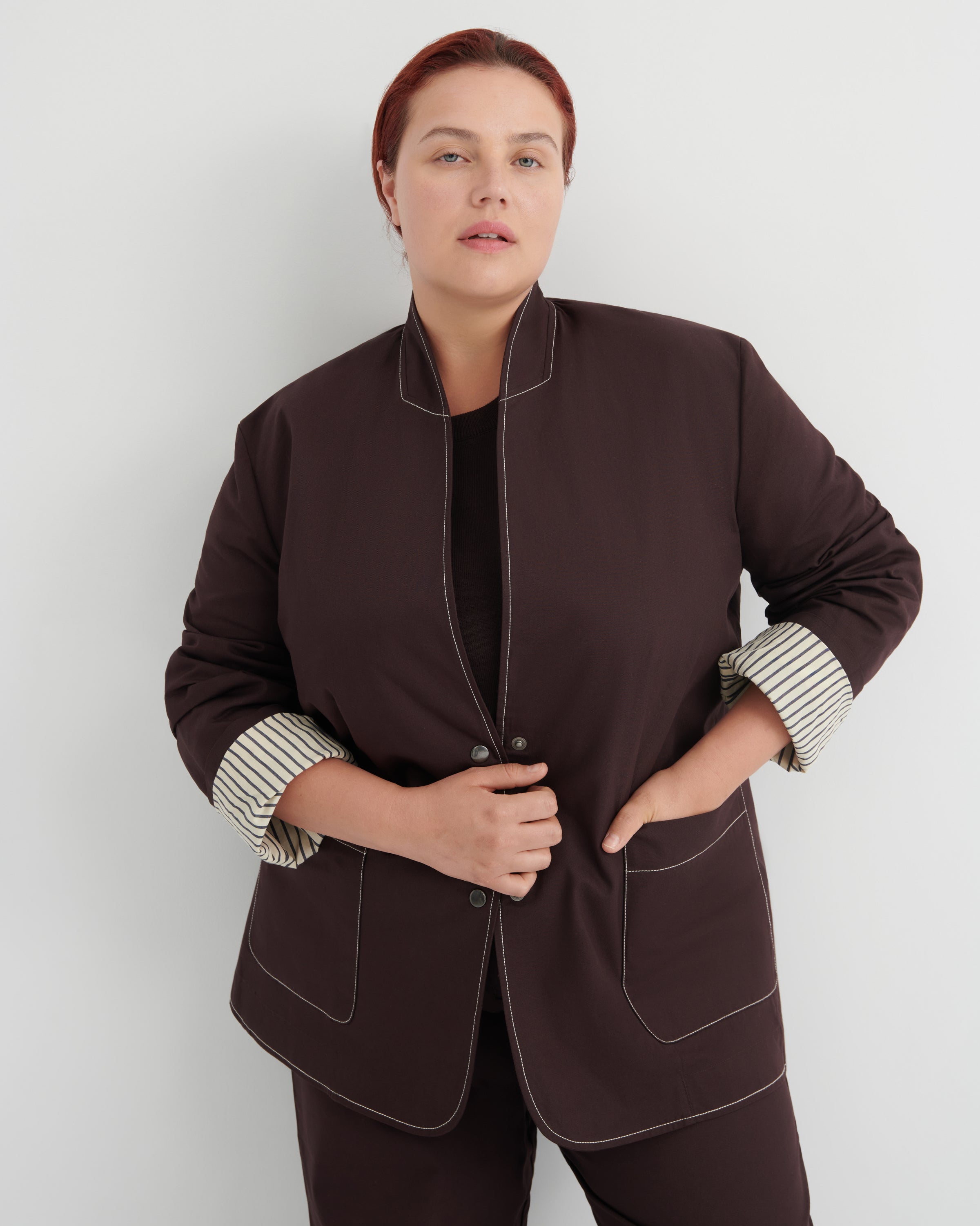 A woman modeling a dark brown jacket with contrasting light topstitching and striped button-up cuffs, paired with matching dark brown trousers against a light gray studio background.